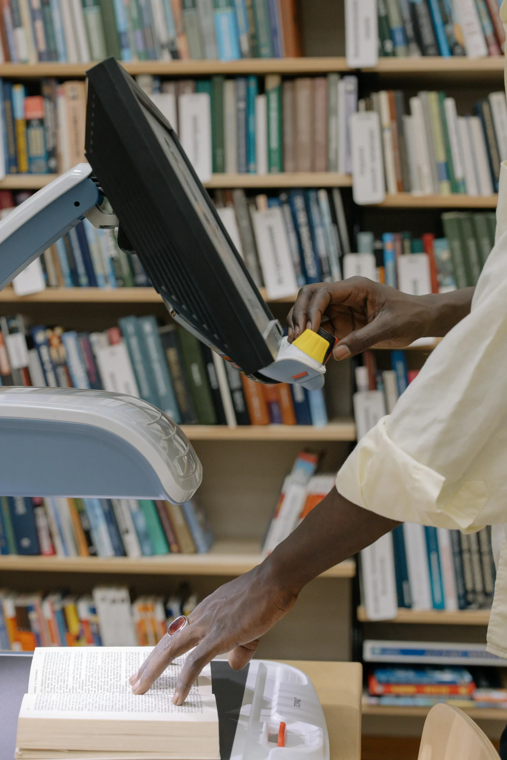 Imagen dentro de una biblioteca que muestra a un hombre buscar información en un monitor, mientras consulta algo en un libro físico